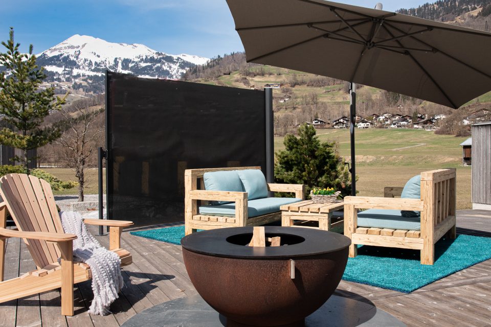 Gartensituation mit Sonnenschirm und einer Lounge aus Massivholz einem Rasenteppich Canada Chairs und eier Feuerkugel sowie einer ombra Wind- und Sichtschutzwand